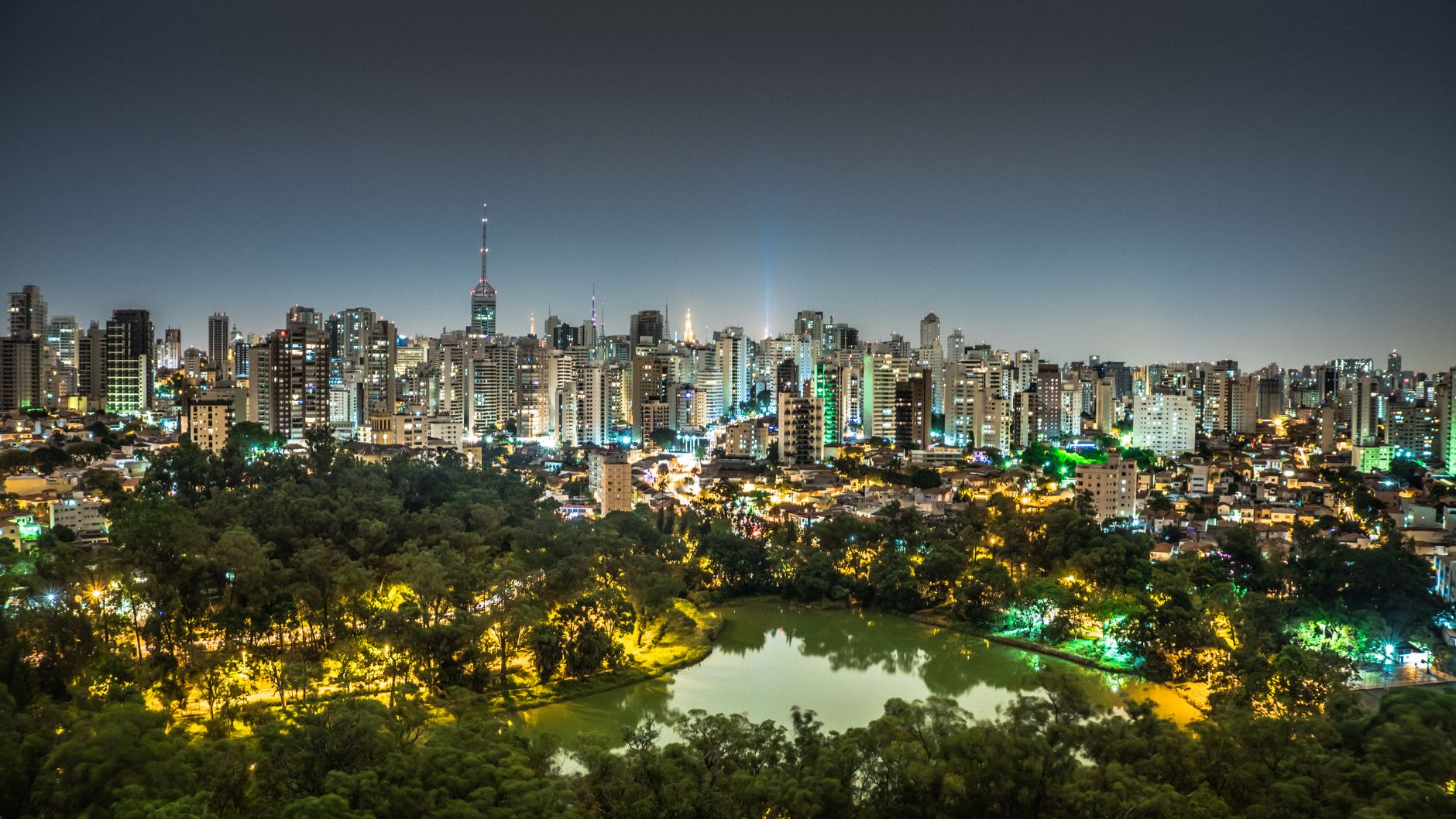 Night view of São Paulo's illuminated skyline with a park and lake in the foreground.