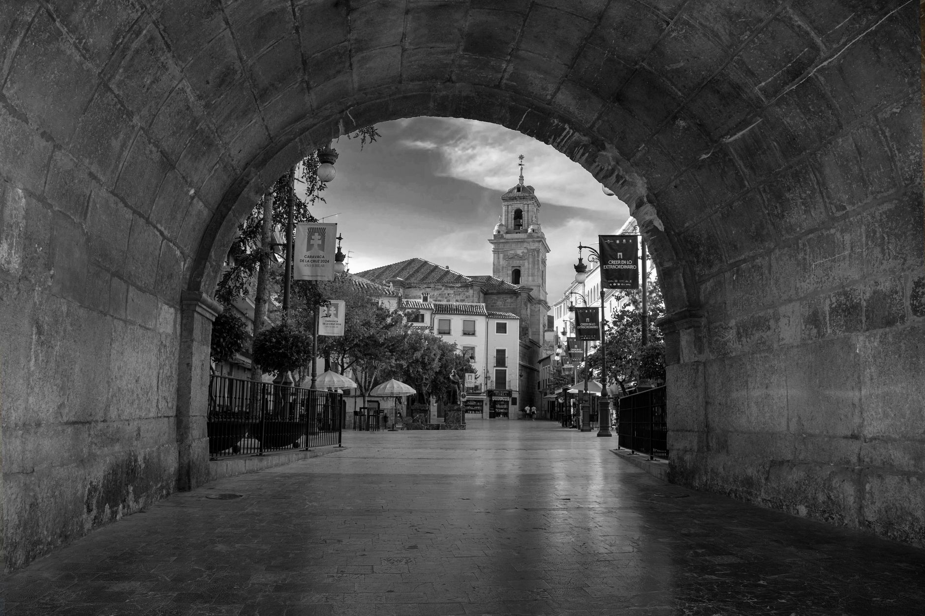 Black and white view through a stone archway leading to a historic Spanish plaza.