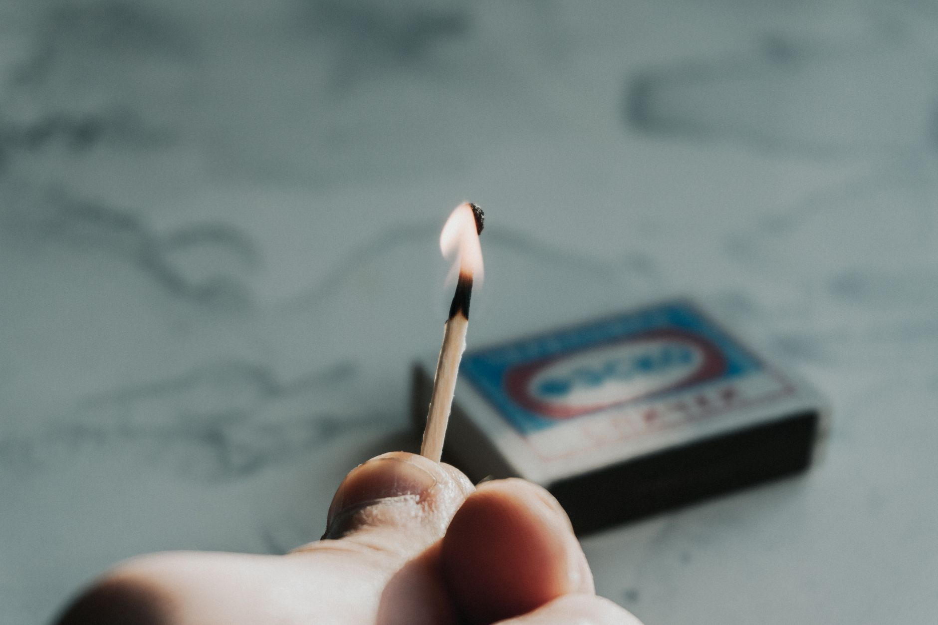 A detailed close-up of a hand holding a burning matchstick against a soft focus background.