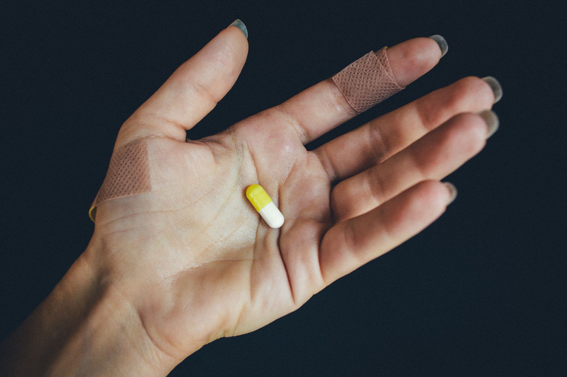 Close-up of a hand holding a yellow capsule with adhesive bandages on fingers.