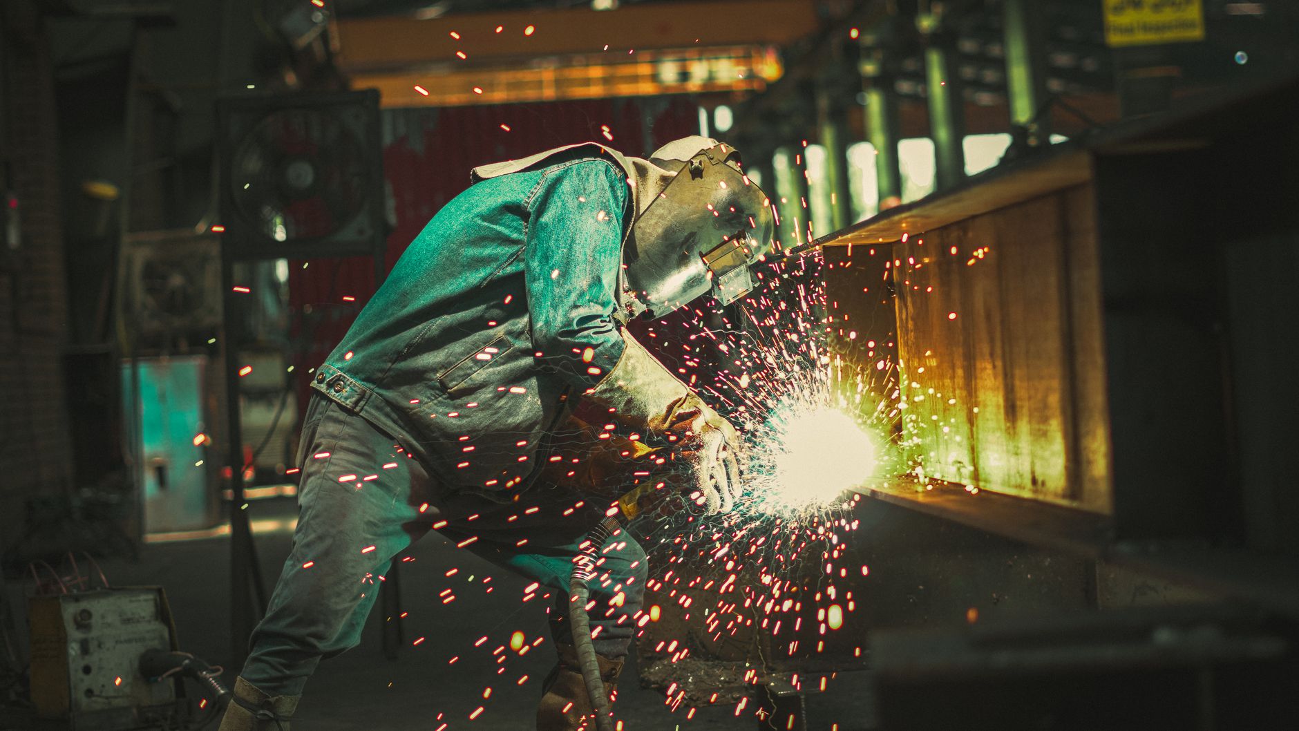 A focused welder wearing protective gear as bright sparks fly in a factory setting.
